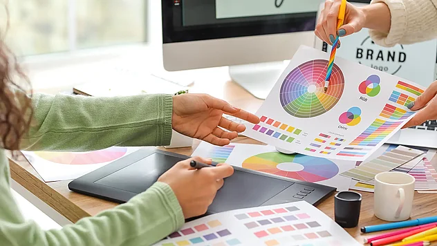 Zwei Personen entwickeln ein Corporate Design mit Farbfächern und Farbmustern am Schreibtisch, im Hintergrund ist ein Bildschirm mit Brand‑Logo zu sehen.