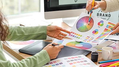 Zwei Personen entwickeln ein Corporate Design mit Farbfächern und Farbmustern am Schreibtisch, im Hintergrund ist ein Bildschirm mit Brand‑Logo zu sehen.